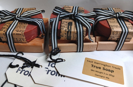 Three bars of red goat milk soap wrapped in brown paper with black and white striped ribbons, on red alder wooden Soap Dishes with gift tags.
