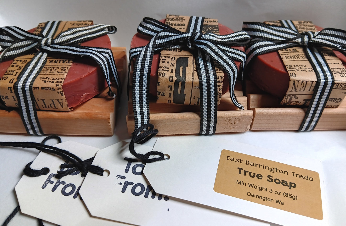 Three bars of red goat milk soap wrapped in brown paper with black and white striped ribbons, on red alder wooden Soap Dishes with gift tags.